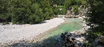 a river with a rocky shore with trees and a house at Apartma Trebušca in Dolenja Trebuša