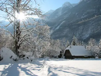 a snow covered cabin with a tree and mountains at Pristava Lepena Village in Lepena