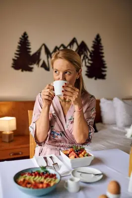 a woman sitting at a table drinking a cup of coffee at Pohorje Village Wellbeing Resort - Wellness & Spa Hotel Bolfenk in Pohorje